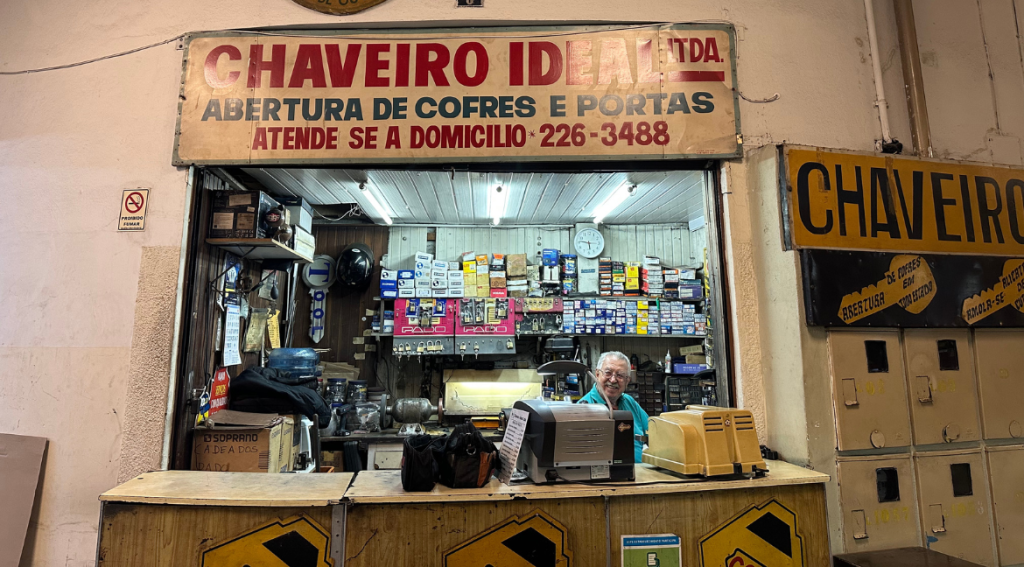 Fotografia digital de Carlos. Ele é idoso, tem cabelos brancos, usa óculos e uma camisa verde e sorri. Está sentado atrás do balcão de sua loja, que tem muitas ferramentas nas paredes. Acima dele, há uma placa de identificação da loja.