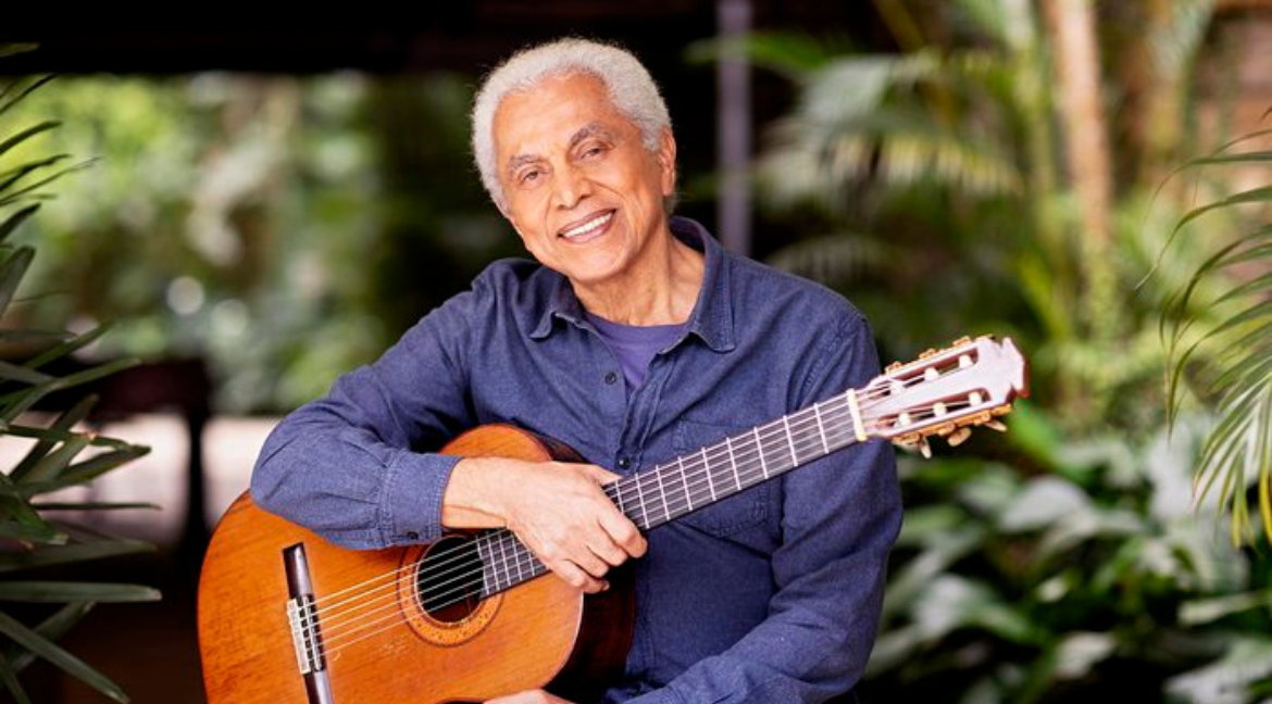 Paulinho da viola é um homem negro, idoso, de cabelos brancos. Na fotos, ele usa camisa azul, segura um violão e sorri, na frente de várias plantas.