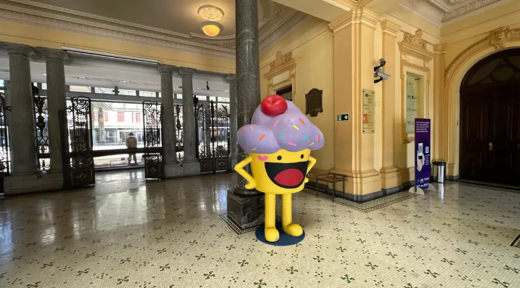 No centro da entrada do Museu de Artes e Ofícios, há um bolinho de cerca de 2 metros de altura. Ele é amarelo, com topo roxo e cereja vermelha no topo.