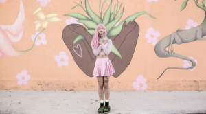 Efe Godoy posa em frente a um mural pintado por ela mesma. Na pintura, folhas saem de um tronco humano com os braços cruzados. Ela é uma mulher transsexual, parda, com tatuagens e cabelo inteiramente rosa. Também usa roupas no mesmo tom do cabelo.