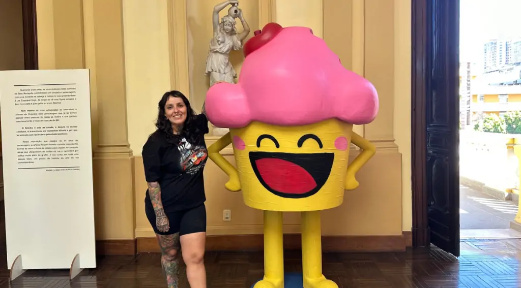 Raquel posa ao lado de um bolinho gigante no museu. Ela é branca, ttuada, tem cabelos lisos e escuros e usa preto. Ele tem cerca de 2 metros, é rosa e amarelo, com uma cereja no topo.
