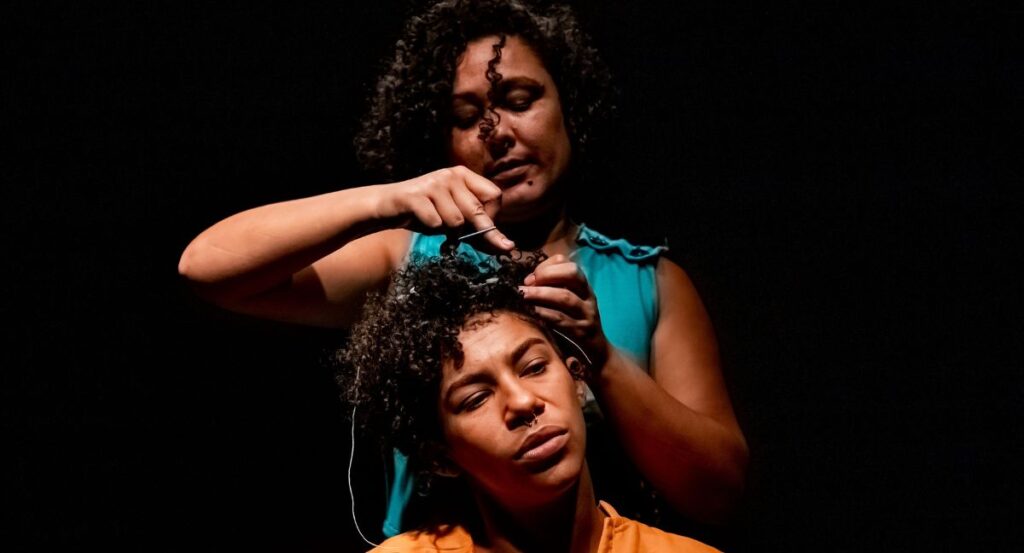 Na foto, de laranja, à frente, Denise Paz, e, ao fundo, Anair Patrícia, em cena de "Querença", apresentada no FIT BH (Denise dos Santos/Renca Produções/Divulgação)